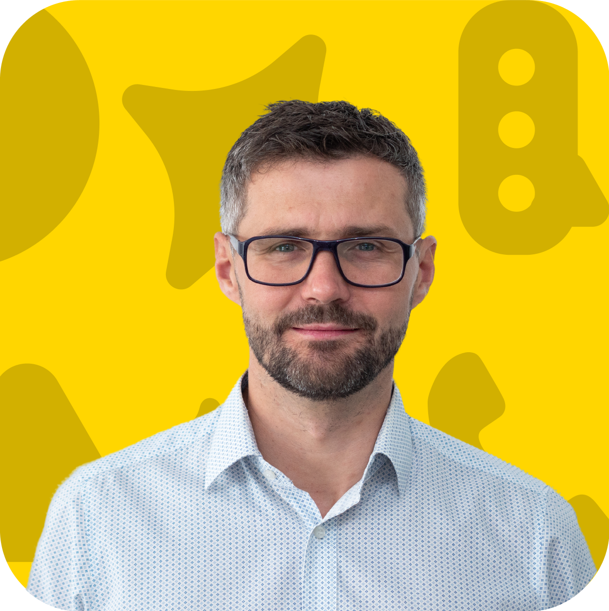 Portrét muže s krátkými hnědými vlasy, bradkou a brýlemi, oblečeného v bílé košili s jemným vzorem. Má neutrální výraz a dívá se přímo do kamery. Pozadí je žluté s abstraktními tvary